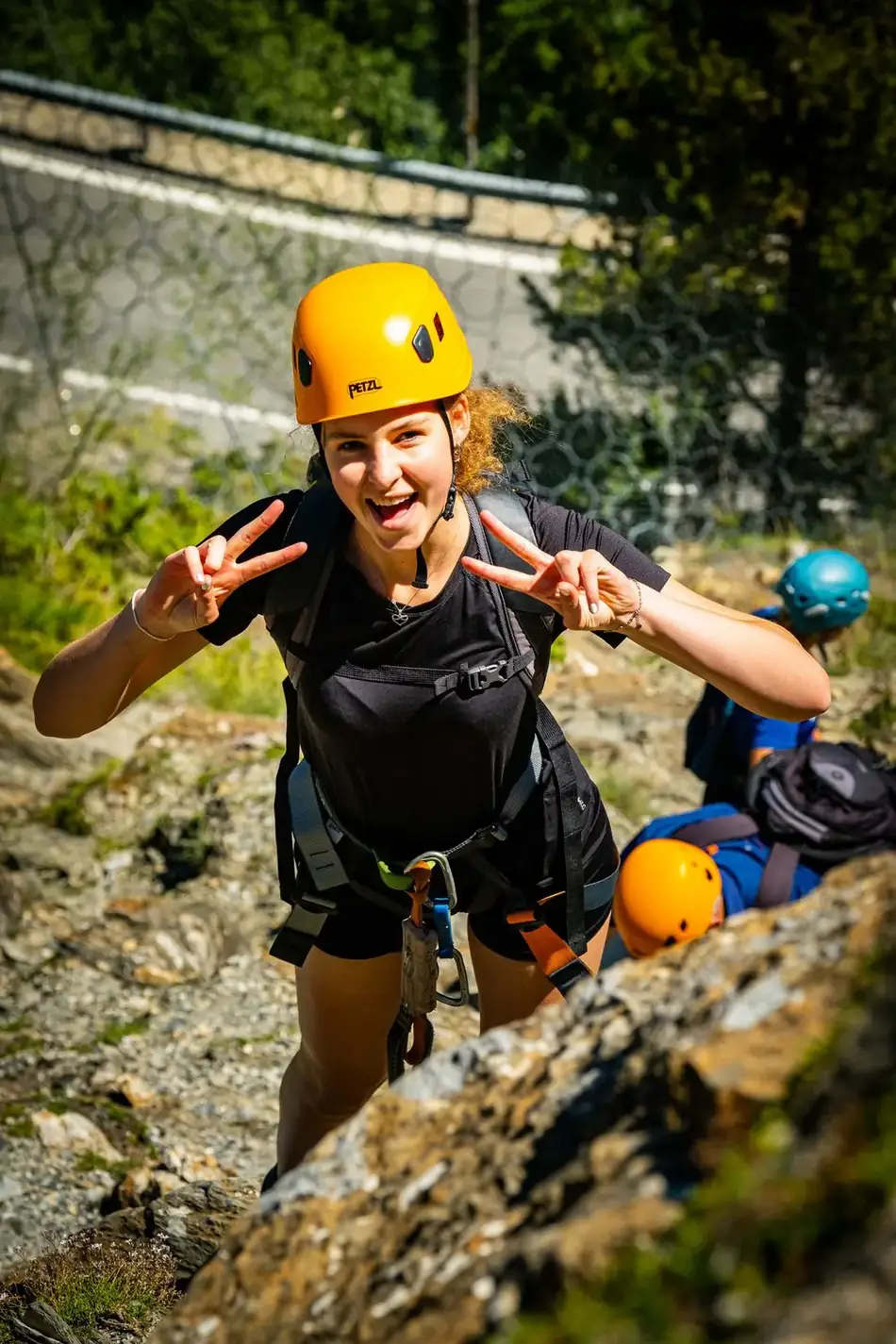 Twee mensen vastgeklikt en lachend op een via ferrata route in Andorra, één maakt een vredesteken